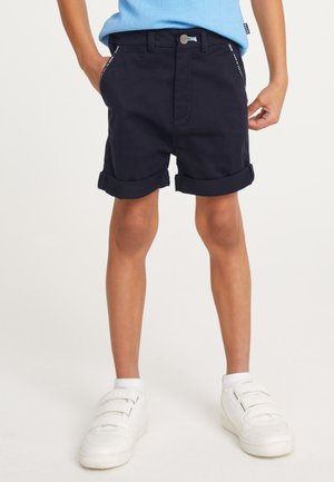 Bambino in piedi con pantaloncini blu navy risvoltati, sneaker bianche con chiusura a velcro e maglietta azzurra, con le mani nelle tasche.