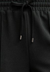 Zwarte trainingsbroek met een gematteerde afwerking, voorzien van een geplooide tailleband en een verstelbaar trekkoord met metalen uiteinden.