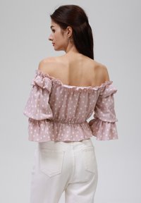 Blouse rose décolletée avec des manches à volants et des pois blancs, cintrée à la taille. Associée à un pantalon blanc taille haute.