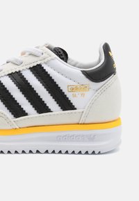 Baskets blanches à rayures noires, en cuir et tissu ; avec accents jaunes et semelle extérieure texturée à bord dentelé.