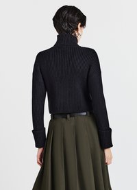 Maglione nero a coste con collo alto e maniche lunghe, con polsini allungati. Abbinato a una gonna plissettata verde oliva.