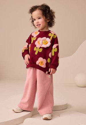 Småbarn med krøllet hår iført en mørkerød sweatshirt med smilende blomster og lyserøde bukser, stående på en platform med abstrakt form.