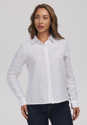 Kobieta w białej koszuli z długim rękawem zapinanej na guziki i ciemnych dżinsach, stojąca z jedną ręką na biodrze, lekko patrząca na bok na szarym tle.