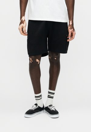 Bărbat purtând pantaloni scurți cargo negri, șosete albe cu dungi negre și pantofi sport negri din pânză, stând sprijinit de un fundal simplu.