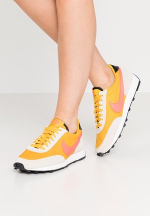 Jambes portant des baskets jaunes et blanches avec des logos swoosh roses, des semelles noires et des lacets jaunes sur un sol gris clair.