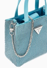 Blå håndtaske prydet med rhinestones, med en sølv kædestrop og et trekantet Guess-logo. Har glatte håndtag og en struktureret form.