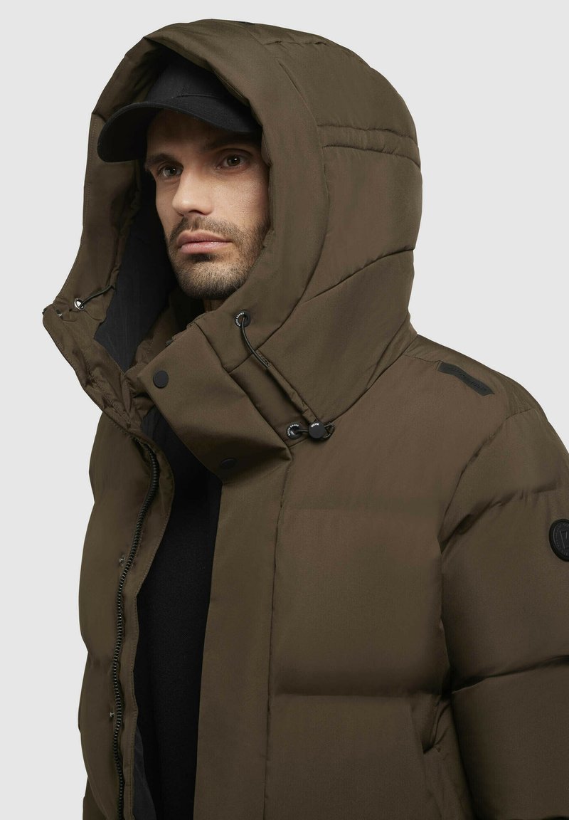 Veste en duvet marron avec une grande capuche, des boutons de fermeture et des cordons de serrage réglables. Elle possède une texture lisse et un patch logo sur la manche.