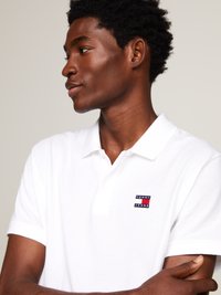 Camisa polo blanca con un tejido texturizado, que presenta un pequeño logo bordado en rojo y azul marino. El cuello y las mangas son cortos.