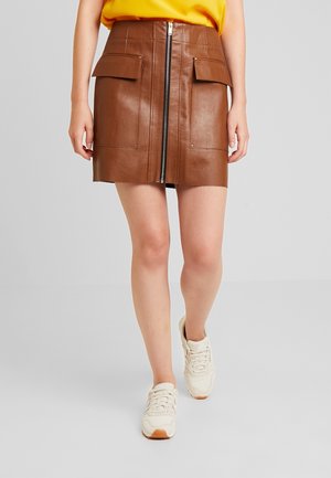 Femme marchant avec une mini-jupe en cuir marron à fermeture éclair avant et poches à rabat, un haut jaune et des baskets blanches sur un fond gris.