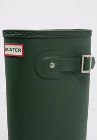Zöld gumicsizma lekerekített tetejével, csat és pánt részletekkel. Egy piros és fehér címke mutatja a "HUNTER" márkát.