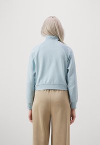 Sweatshirt bleu clair court avec un col haut et des poignets côtelés, présentant un tissu texturé et des manches raglan. Pantalons beige amples.