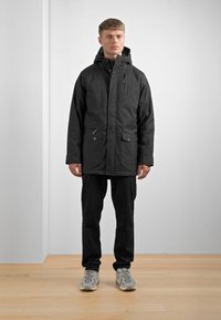 Svart vanntett jakke med frontglidelås, to sidelommer og hette, kombinert med svarte bukser og grå sneakers, på et tregulv.