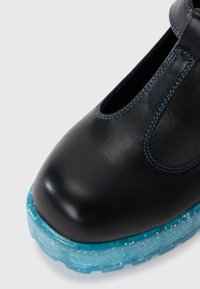 Chaussure en cuir noir avec un bout arrondi et un design découpé, présentant une semelle mouchetée bleu clair et des coutures bleues contrastées.