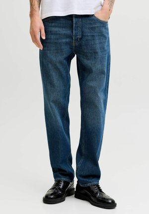 Mann trägt dunkelblaue gerade geschnittene Jeans, schwarze Lederschuhe und ein weißes Hemd mit einer Hand in der Tasche.