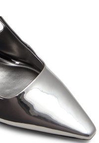 Acercamiento de un zapato de tacón alto brillante de punta en plata con forro interior negro sobre un fondo blanco.