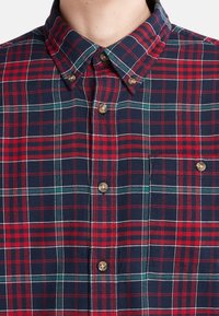 Camicia a quadri con colori rosso, blu navy e verde; colletto button-down; taschino sul petto; e orlo arrotondato con bottoni marroni.