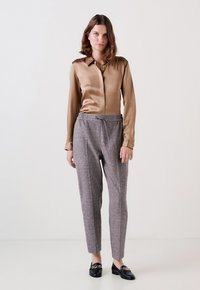 Satin bruine blouse met een kraag, gecombineerd met grijze broek met patroon en elastische tailleband, en zwarte instappers met een kettingaccent.