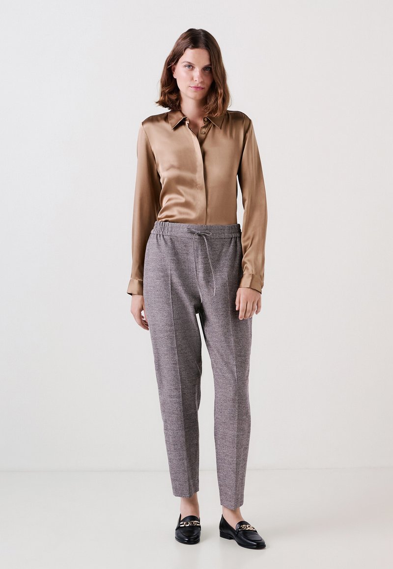 Satin bruine blouse met een kraag, gecombineerd met grijze broek met patroon en elastische tailleband, en zwarte instappers met een kettingaccent.