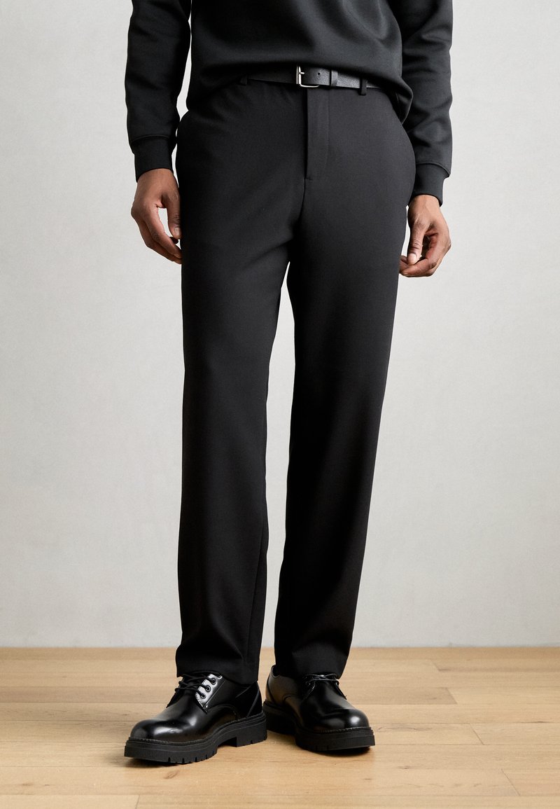 Pantalons noirs en tissu lisse, avec une coupe droite, des passants de ceinture et une taille plate. Associés à des chaussures noires brillantes.