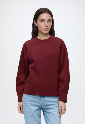Femme aux longs cheveux foncés porte un sweat-shirt bordeaux uni et un jean bleu clair, debout devant un fond blanc.