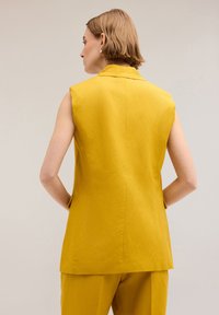 Gelber ärmelloser Blazer mit strukturiertem Kragen, glattem Stoff und Seitentaschen, der eine taillierte Passform und ein minimalistisches Design aufweist.