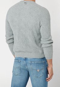 Homme portant un pull en maille texturée gris clair et un jean en denim bleu, montré de dos avec la main droite détendue à ses côtés.