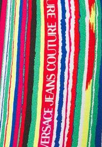 Rayures verticales multicolores avec le texte "VERSACE JEANS COUTURE" en blanc sur une bande rouge, sur un fond en tissu texturé.
