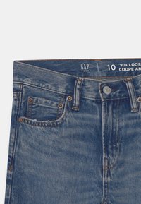 Blauwe spijkerbroek met taille en knoopsluiting, riemlussen, klinknagels op de voorkant en een 'GAP Teen' label dat maat 10 aangeeft in een losse stijl uit de jaren '90.