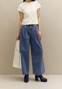 Vit croppad t-shirt med korta ärmar, kombinerad med hög midja, vida blå denimjeans. Svarta klackade ankelboots och en vit tote-väska.