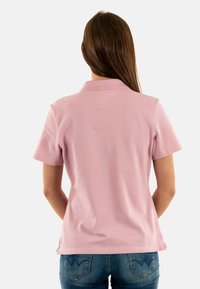 Polo rosa a maniche corte con colletto, realizzata in tessuto liscio, con spacco sul fondo e dettagli di cuciture sottili. Indossata con jeans blu.