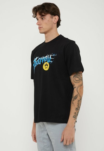 Young man with tattooed arm wearing black graphic t-shirt with blue text and yellow smiley face, standing sideways against plain background.