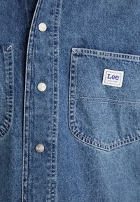 Camicia in denim blu con bottoni a pressione argentati, taschino sul petto e un'etichetta Lee ben visibile con sfondo bianco e testo blu.