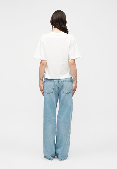 Άτομο με μακριά σκούρα μαλλιά που φοράει απλό λευκό κοντομάνικο μπλουζάκι και ανοιχτό μπλε φαρδιά τζιν, στέκεται γυρισμένο προς τα πίσω σε λευκό φόντο.