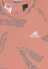 Růžové sportovní tričko s kulatým výstřihem, na přední straně s šedým nápisem a bílým logem Adidas, vyrobené z hladké látky.