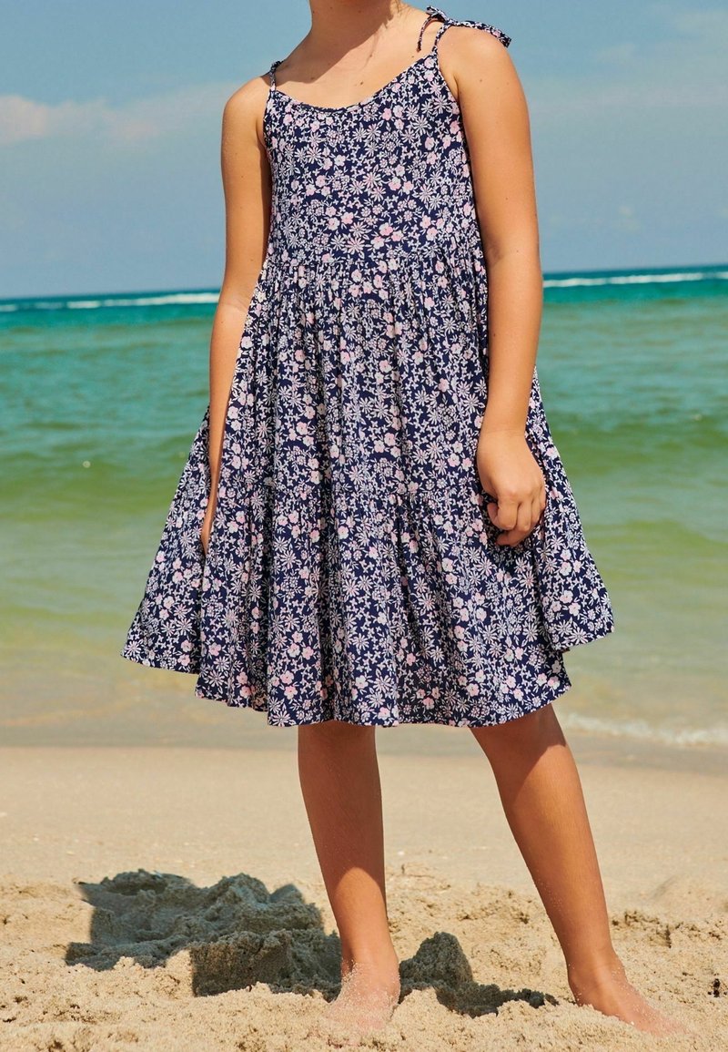 Robe florale bleu marine à fines bretelles, taille froncée et jupe évasée, avec des motifs de fleurs roses et blanches. Portée sur une plage de sable.