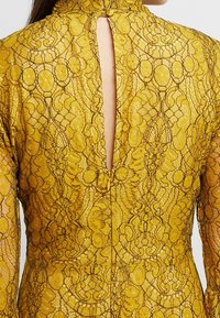 Vestido amarillo de encaje con patrones florales intrincados, cuello alto y una abertura en forma de ojo de cerradura en la parte trasera. Texturizado con un acabado translúcido.