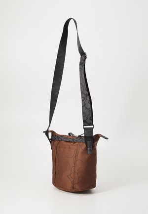 Braune Bucket-Style Crossbody-Tasche mit schwarzen Blumenmustern und einem verstellbaren schwarzen und gemusterten Schulterriemen, dargestellt vor weißem Hintergrund.