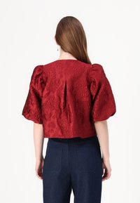 Red textured blouse with puffed sleeves, featuring floral patterns and a back pleat, paired with dark blue denim trousers.