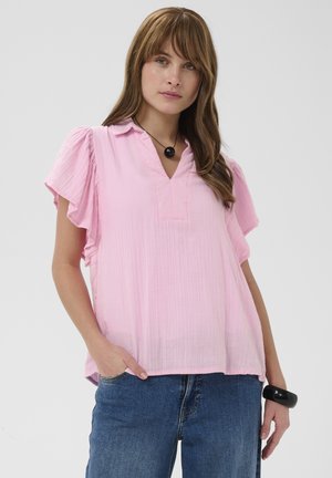 Kobieta z długimi brązowymi włosami, ubrana w jasnoróżową bluzkę z krótkim rękawem, niebieskie dżinsy, czarny naszyjnik i czarną bransoletkę, stojąca na jednolitym tle.