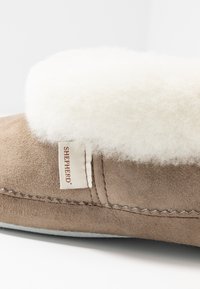 Suede slipper in light brown with a fluffy white sheepskin collar, featuring stitched detailing and a branded tag on the side.