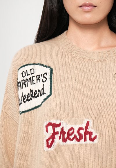 Beige strikkegenser med to lapper: den ene har teksten "OLD FARMER'S WEEKEND" i svart på hvit bakgrunn, den andre "Fresh" i rød skrift.