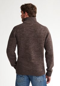 Man die met zijn rug naar de camera staat, gekleed in een bruine hoge hals trui en een spijkerbroek tegen een plain witte achtergrond.
