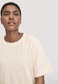 Erwachsene Person mit kurzem, lockigem Haar, die ein einfarbiges, off-white T-Shirt trägt, vor einem hellgrauen Hintergrund steht und leicht nach oben schaut.