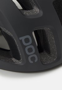 Casco de ciclismo negro con aberturas de ventilación, superficie texturizada y un sutil patrón similar al carbono. Presenta un logo en gris de "POC" en un lateral.