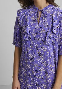 Floral dress in purple fabric featuring ruffles along the shoulders and neckline, with black, pink, and yellow flower patterns.