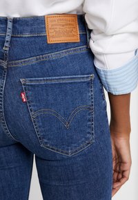 Jeans de mezclilla azul oscuro con un parche de cuero marrón, costuras doradas y un distintivo detalle en el bolsillo trasero en forma de corazón.
