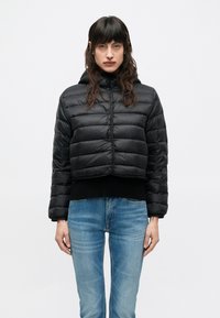 Μαύρο κοντό μπουφάν puffer με οριζόντια ραφή, φερμουάρ μπροστά και κουκούλα. Φορεμένο πάνω από μαύρο πουλόβερ και μπλε τζιν.