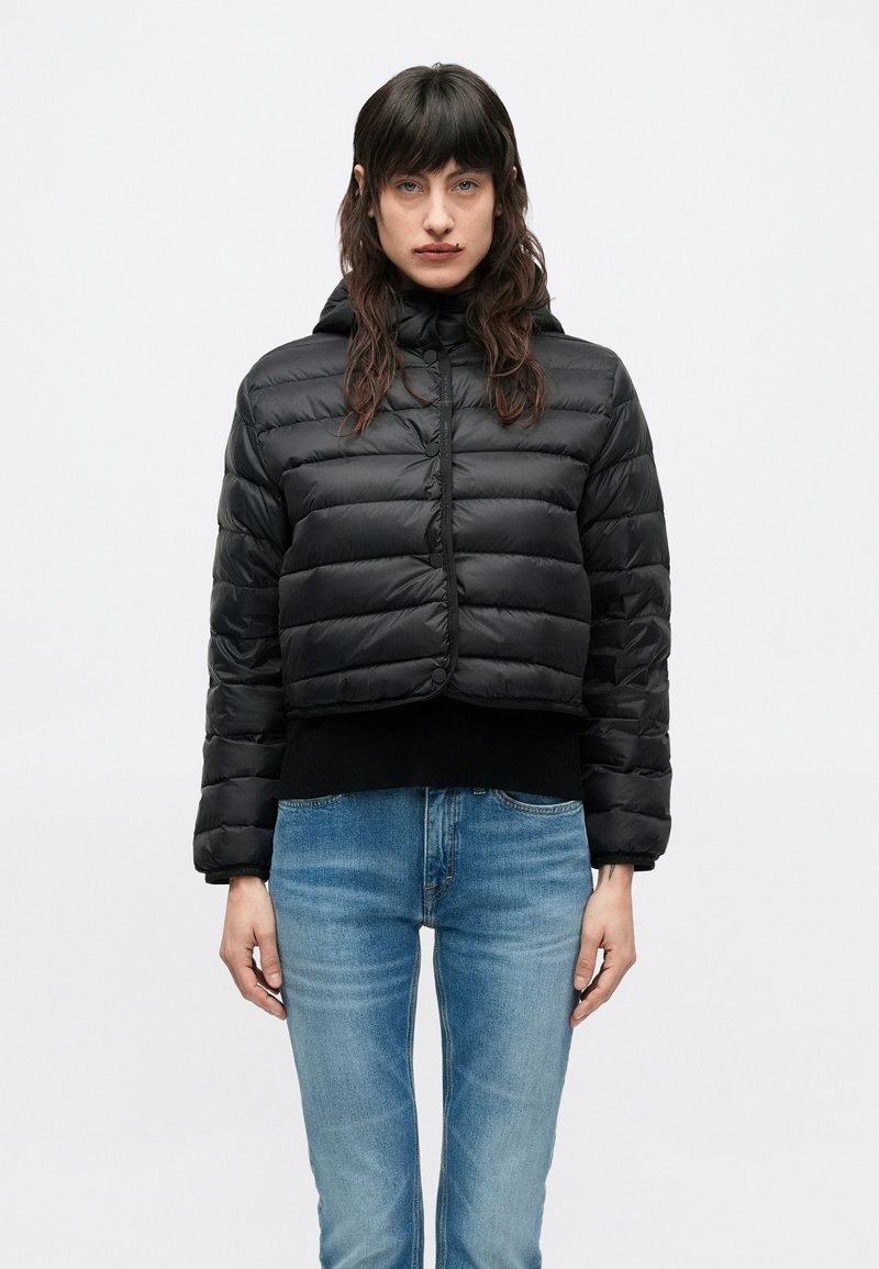 Μαύρο κοντό μπουφάν puffer με οριζόντια ραφή, φερμουάρ μπροστά και κουκούλα. Φορεμένο πάνω από μαύρο πουλόβερ και μπλε τζιν.