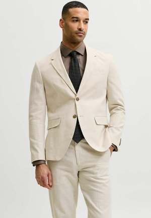 Uomo con un abito beige, camicia marrone scuro e cravatta nera a fantasia, in piedi con una mano in tasca, che guarda di lato.