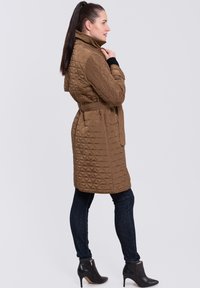 Abrigo trench de color marrón acolchado con cuello alto, cinturón y dos bolsillos laterales. Llevado sobre jeans oscuros y botas negras de tobillo. Tejido suave y texturizado.